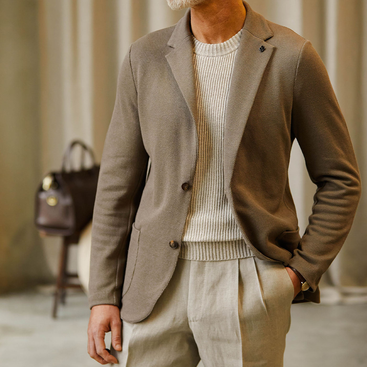 A person wearing a stylish Gran Sasso taupe brown cotton linen knitted blazer over a ribbed white sweater paired with light grey trousers, posing with one hand in their pocket. A brown leather bag is visible in the background.