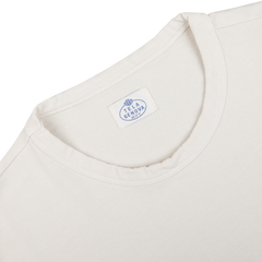 A close-up of an ecru white long-sleeve t-shirt collar made from heavy organic cotton, featuring a white tag that displays the brand name "Tela Genova" in blue lettering.