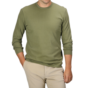 A person is standing with one hand in their pocket, dressed in a Tela Genova Grass Green Heavy Organic Cotton LS T-Shirt and beige pants. Their face is not visible.