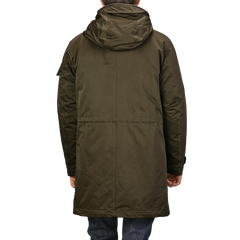 The image features the back view of a person wearing a Ten C Green Technical Rainstorm Parka, made from waxed material, paired with blue jeans against a plain background.