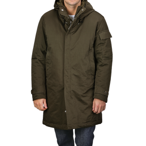 A person is wearing a knee-length Green Technical Rainstorm Parka by Ten C, crafted from waxed material, featuring a hood and snap closures.