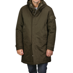 A person is wearing a knee-length Green Technical Rainstorm Parka by Ten C, crafted from waxed material, featuring a hood and snap closures.