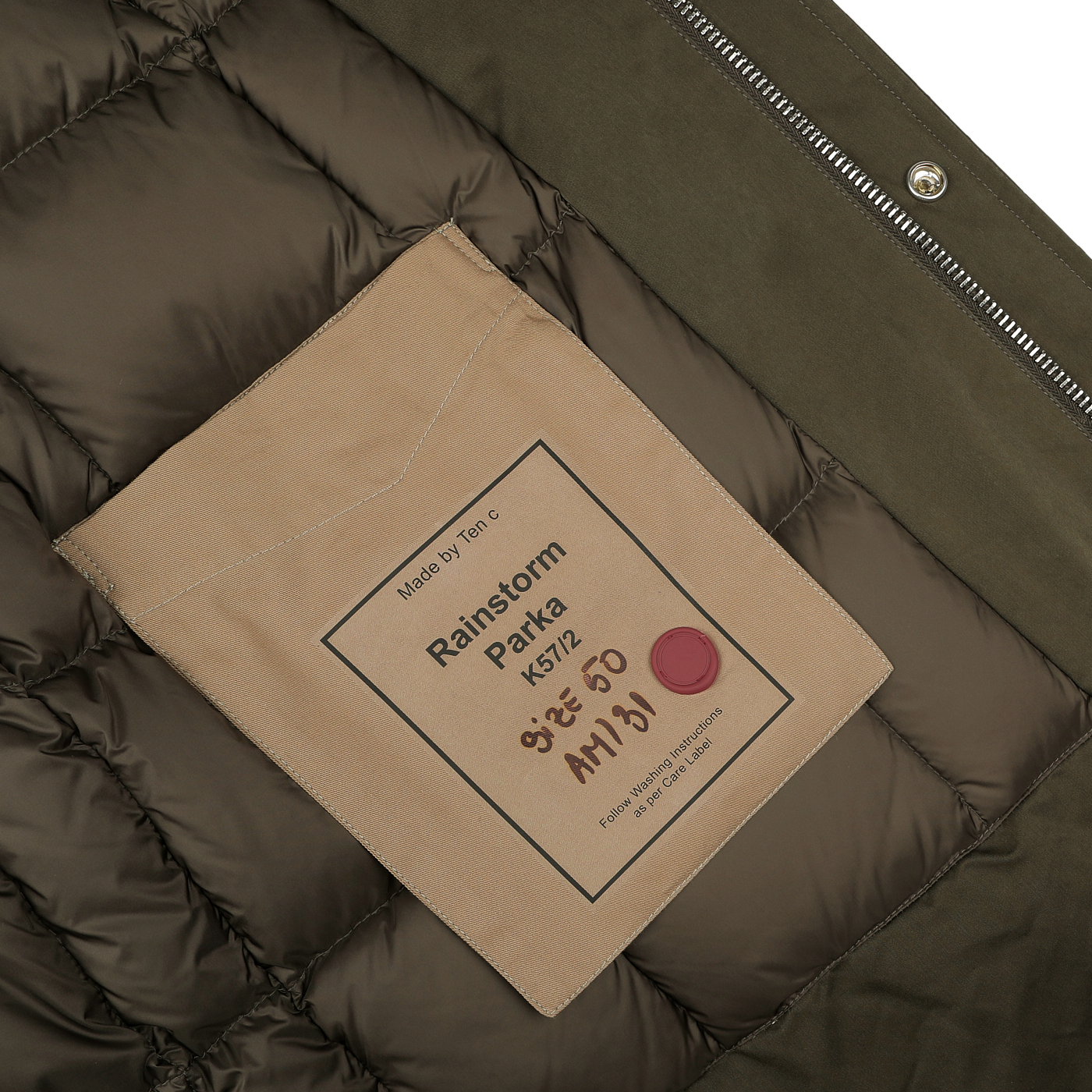 Close-up of a dark green and brown interior of the Ten C Green Technical Rainstorm Parka, crafted from waxed material. It features a beige pocket, a striking red button accent, and a label with care instructions that underscore its durable polyester and polyamide blend.