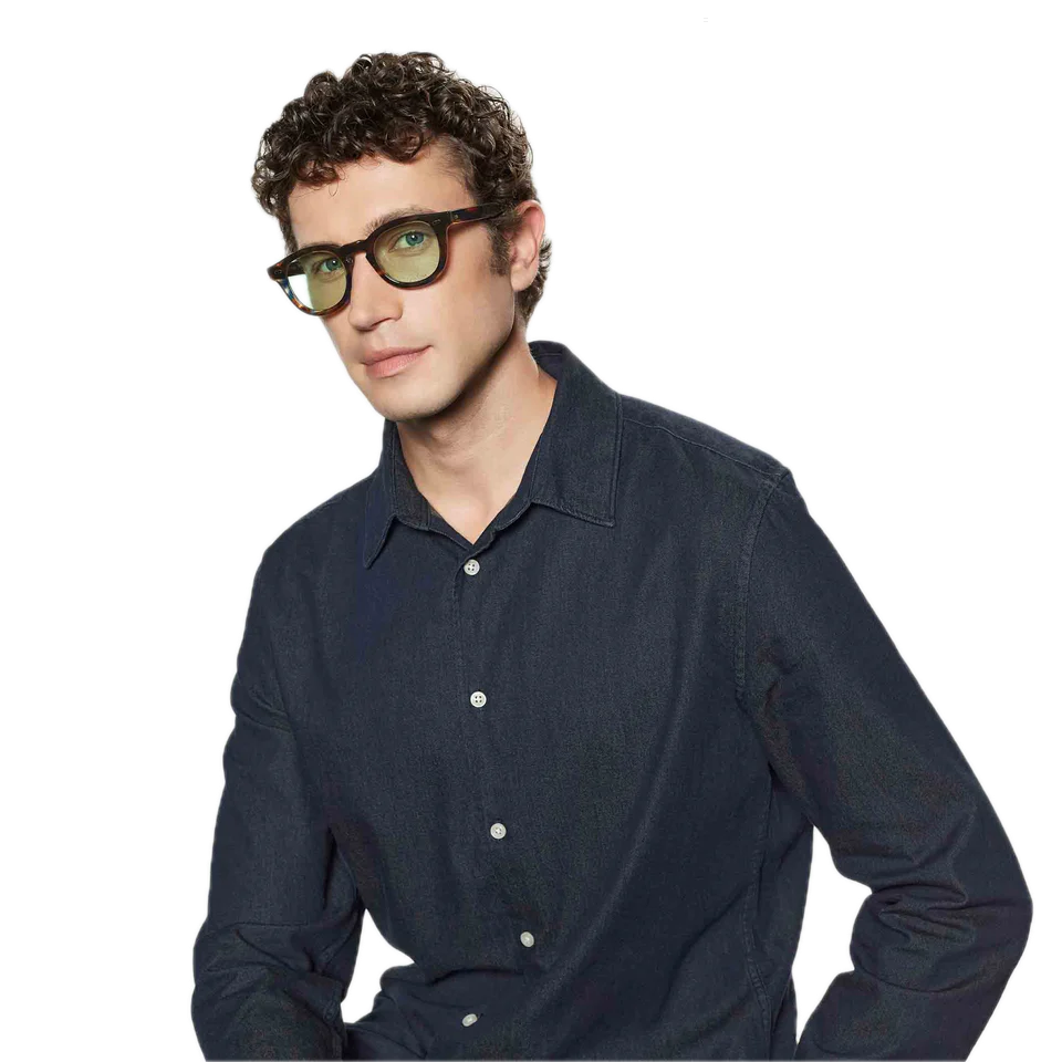 Young man with curly hair and squared frame glasses, wearing a dark blue shirt, stands against a blank background wearing The Bespoke Dudes Donegal River Light Green Sunglasses 49mm.