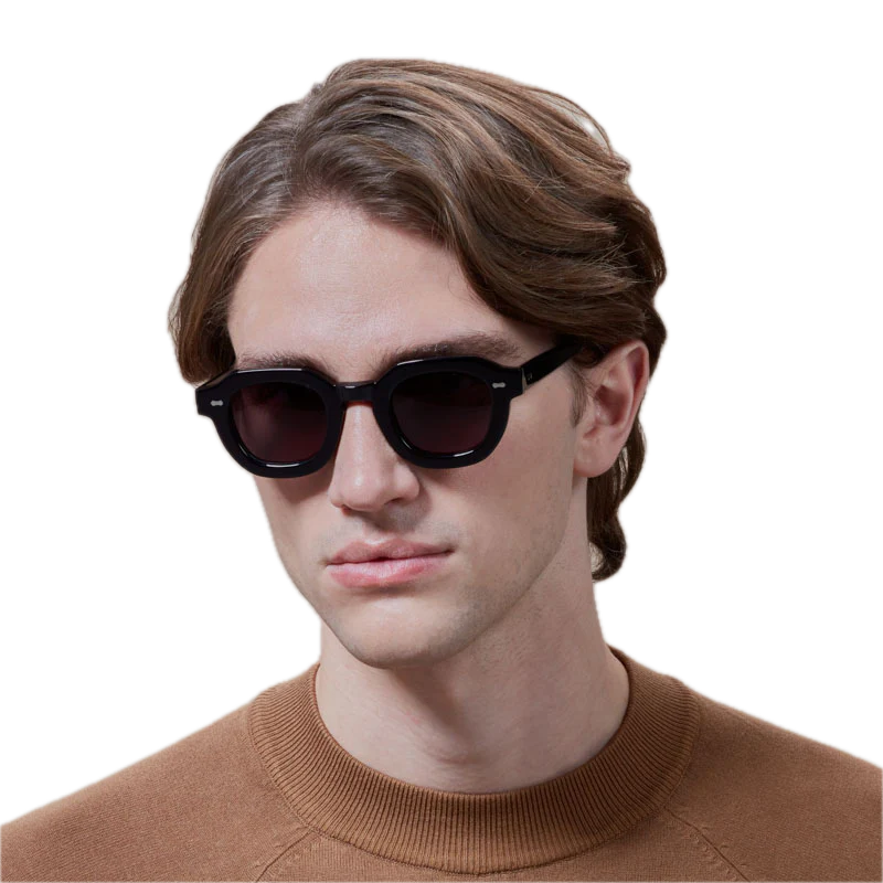 A young man wearing The Bespoke Dudes Juta Eco Black Grey Lenses 46mm sunglasses and a brown sweater, with medium-length wavy hair, against a black background.