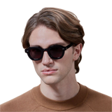 A young man wearing The Bespoke Dudes Juta Eco Black Grey Lenses 46mm sunglasses and a brown sweater, with medium-length wavy hair, against a black background.