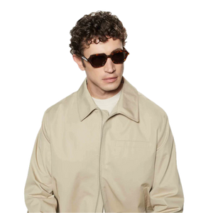 Young man with curly hair wearing square-shaped sunglasses from The Bespoke Dudes, standing against a black background.