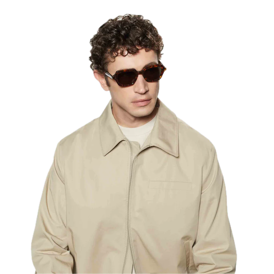 Young man with curly hair wearing square-shaped sunglasses from The Bespoke Dudes, standing against a black background.