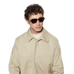 Young man with curly hair wearing square-shaped sunglasses from The Bespoke Dudes, standing against a black background.