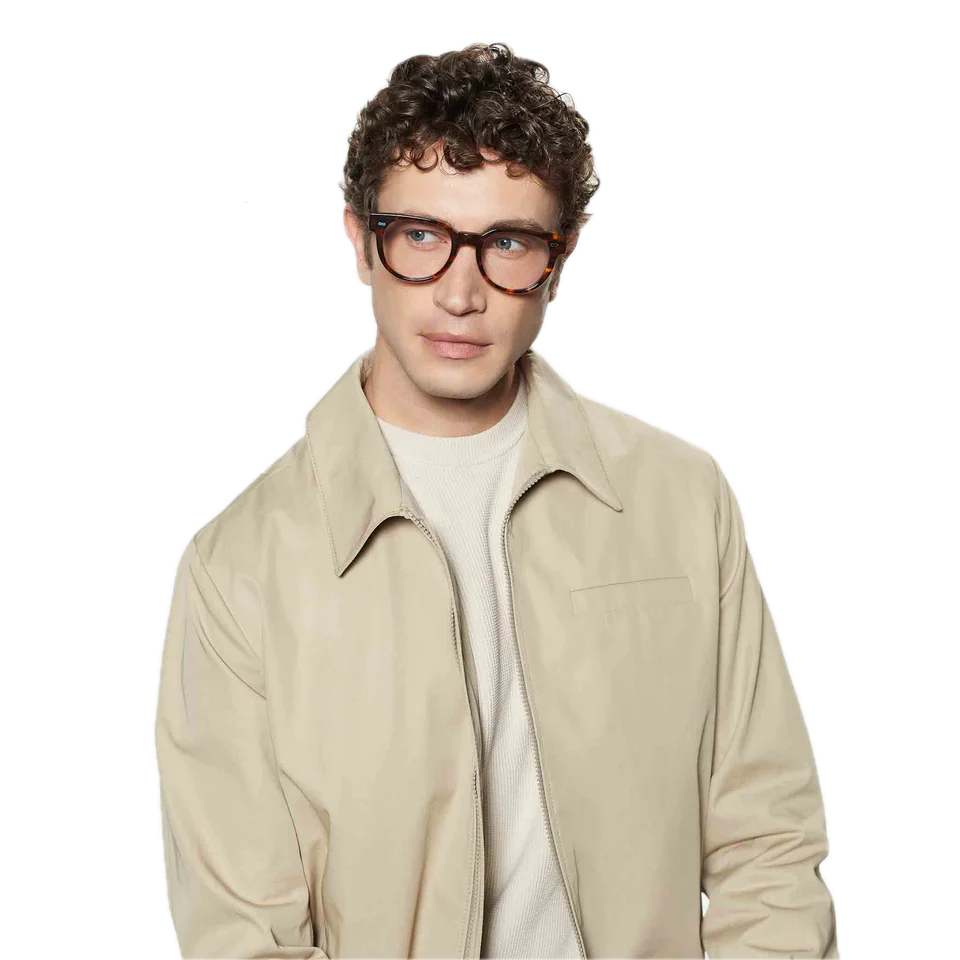 A young man with curly hair wearing sunglasses and a beige jacket over a white shirt, standing against a black background, and wearing The Bespoke Dudes Palm Eco Spotted Havana Optical 51mm glasses.