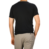 Rear view of a person standing, wearing The White Briefs' Black Pima Cotton Earth T-Shirt made from organic pima cotton and beige pants against a plain background.