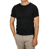 A person is standing, wearing a Black Pima Cotton Earth T-Shirt from The White Briefs and beige pants, with their hands resting in their pockets.