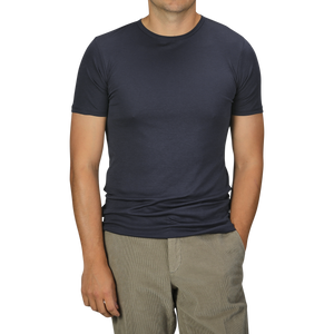 A man in a Parisian Night Pima Cotton Earth T-Shirt by The White Briefs and beige pants stands with his hands partially in his pockets against a gray background.