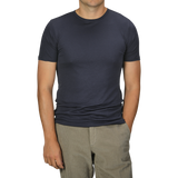 A man in a Parisian Night Pima Cotton Earth T-Shirt by The White Briefs and beige pants stands with his hands partially in his pockets against a gray background.