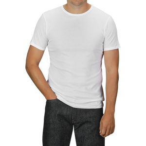 A person wearing a White Pima Cotton Earth T-Shirt by The White Briefs and slim fit dark jeans stands with their left hand in their pocket. The background is a neutral grey.