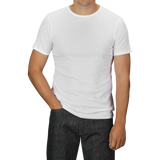 A person wearing a White Pima Cotton Earth T-Shirt by The White Briefs and slim fit dark jeans stands with their left hand in their pocket. The background is a neutral grey.