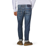 A person is standing with their back to the camera, wearing Tramarossa's Washed Blue Loro Piana Cotton Michealangelo Jeans and brown shoes. The jeans, made from super stretch material, are slightly cuffed at the bottom.