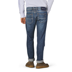 A person is standing with their back to the camera, wearing Tramarossa's Washed Blue Loro Piana Cotton Michealangelo Jeans and brown shoes. The jeans, made from super stretch material, are slightly cuffed at the bottom.