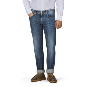 A person wearing a white button-up shirt, Tramarossa's Washed Blue Loro Piana Cotton Michealangelo Jeans with rolled cuffs, and brown shoes stands with one hand in their pocket against a plain backdrop.