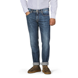 A person wearing a white button-up shirt, Tramarossa's Washed Blue Loro Piana Cotton Michealangelo Jeans with rolled cuffs, and brown shoes stands with one hand in their pocket against a plain backdrop.