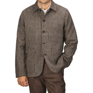 A person wearing a Brown Melange Harris Tweed Bakers C Jacket by Universal Works and brown trousers stands against a plain background. Their right hand is in their pocket, and their head is not visible in the frame.