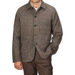 A person wearing a Brown Melange Harris Tweed Bakers C Jacket by Universal Works and brown trousers stands against a plain background. Their right hand is in their pocket, and their head is not visible in the frame.