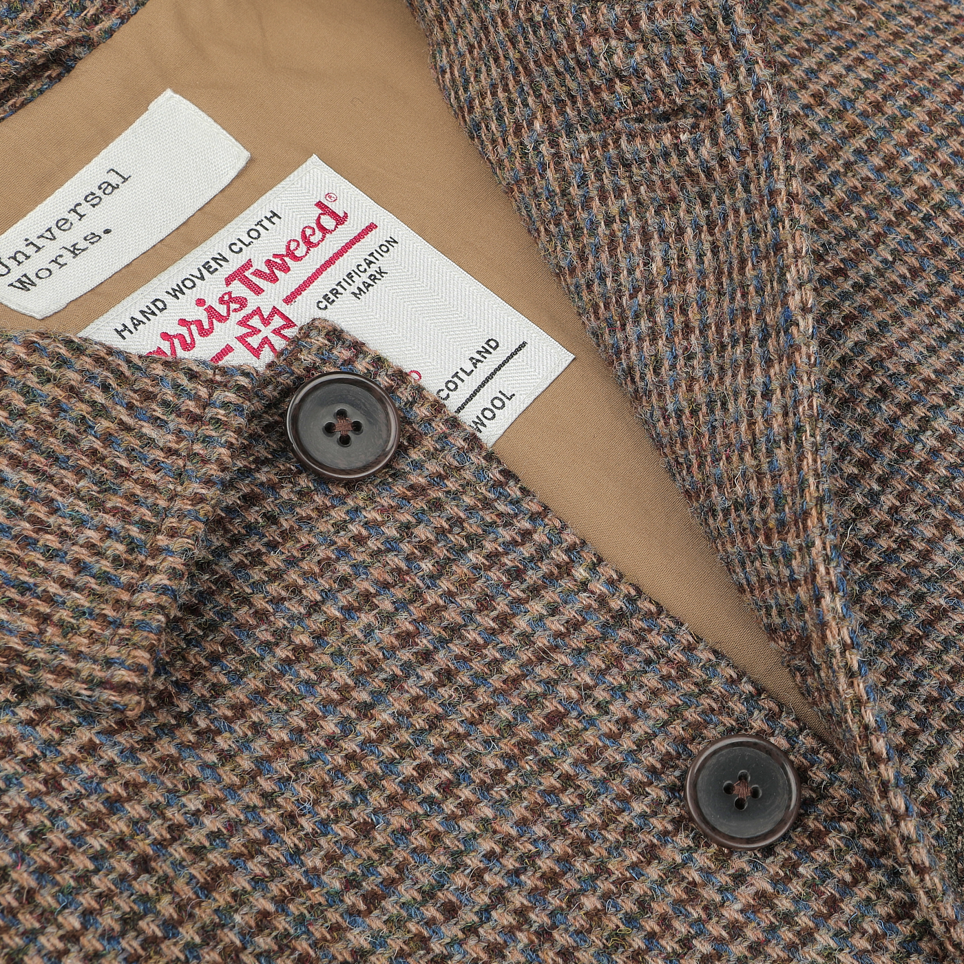 Close-up of a Brown Melange Harris Tweed Bakers C Jacket from Universal Works, showcasing its intricate brown and blue patterns. The jacket features two black buttons and two labels: one reading "Universal Works" and the other "Harris Tweed.