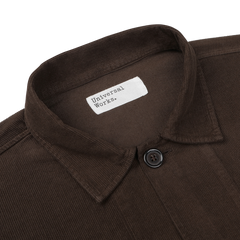 A close-up image of a Dark Brown Fine Corduroy Bakers Overshirt by Universal Works, showcasing a single button and the white label with the brand name.