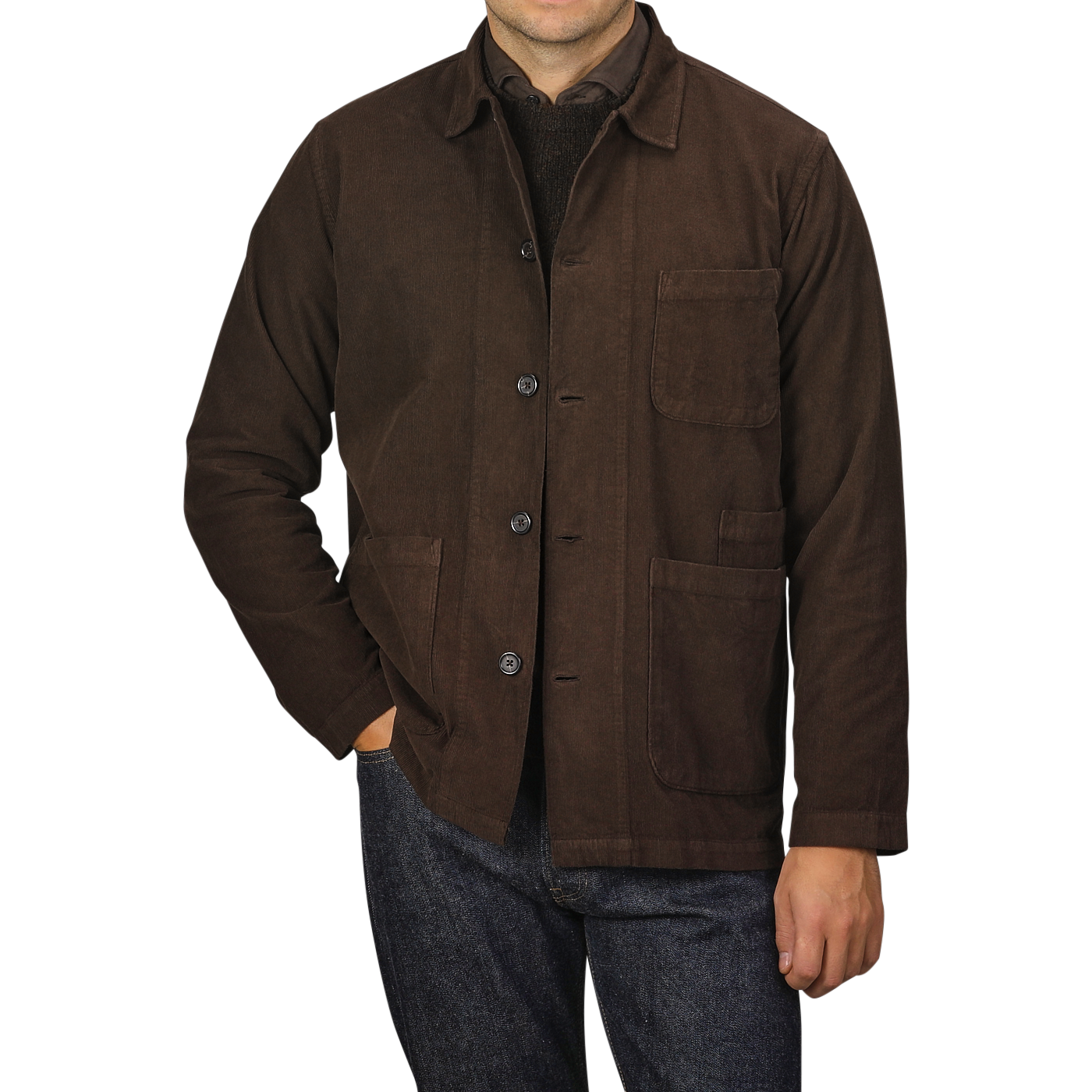 A person is wearing a Universal Works Dark Brown Fine Corduroy Bakers Overshirt over a dark shirt, with their hand casually placed in one of the front pockets of the overshirt. They are also sporting blue jeans, exuding effortless style.