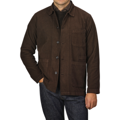 A person is wearing a Universal Works Dark Brown Fine Corduroy Bakers Overshirt over a dark shirt, with their hand casually placed in one of the front pockets of the overshirt. They are also sporting blue jeans, exuding effortless style.