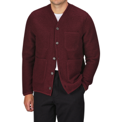 A person wearing the Deep Red Wool Fleece Cardigan by Universal Works over a white shirt, paired with black trousers. The stylish piece, featuring two front pockets, evokes the timeless elegance characteristic of Universal Works.