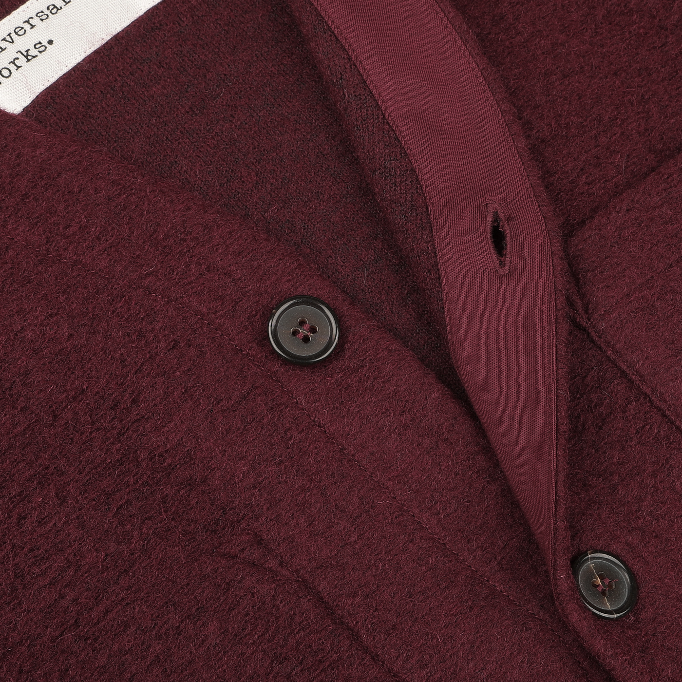 Close-up of a Deep Red Wool Fleece Cardigan by Universal Works, highlighting the fabric texture, buttons, and a partially visible white clothing label.