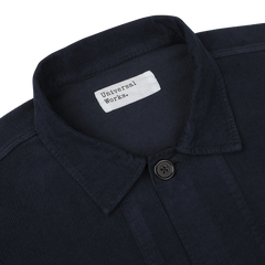 Close-up of a navy fine corduroy Bakers overshirt with a buttoned collar and a white label that reads "Universal Works.