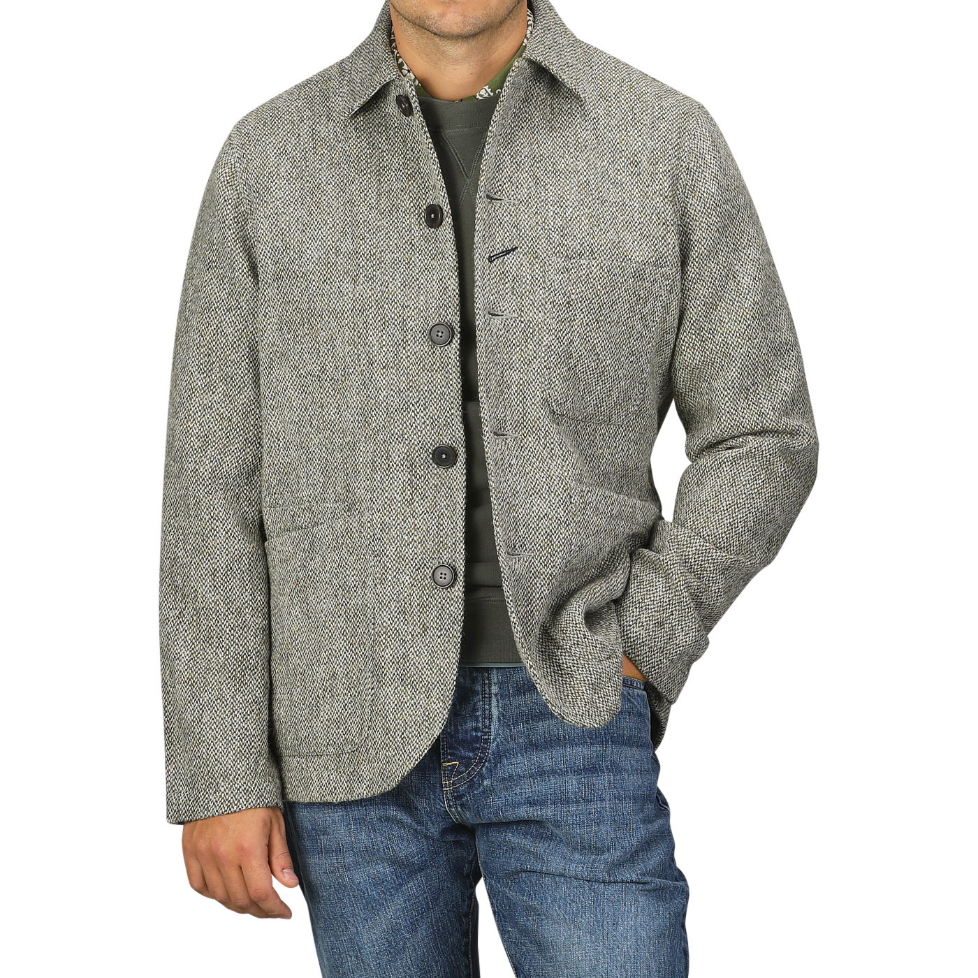 A man wearing an Olive Melange Harris Tweed Bakers C Jacket from Universal Works over a green shirt and blue jeans poses with one hand in his pocket against a plain background.