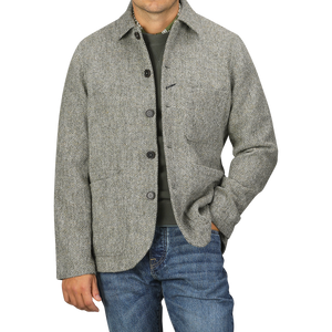 A man wearing an Olive Melange Harris Tweed Bakers C Jacket from Universal Works over a green shirt and blue jeans poses with one hand in his pocket against a plain background.