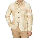 A man wearing a sand beige camo cotton Universal Works bakers jacket and tan pants.