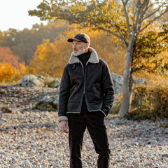 A man in a dark brown leather Alim flight jacket by Werner Christ and a cap stands on a rocky path, surrounded by autumn trees and foliage in the background.