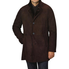 A person wearing a Werner Christ Dark Brown Suede Leather Blake Coat with three buttons is standing with one hand in their pocket against a plain background. The coat features a high collar and reaches mid-thigh, making it perfect for winter weather. The person's face is not visible.