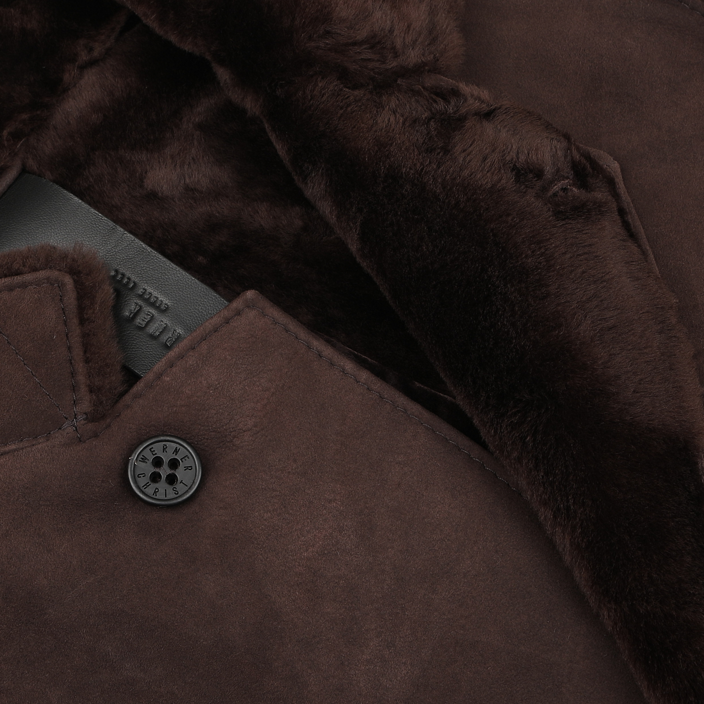 Close-up view of the Werner Christ Dark Brown Suede Leather Blake Coat, featuring a fur-lined interior, a button, and part of a leather strap visible—perfect for winter weather.