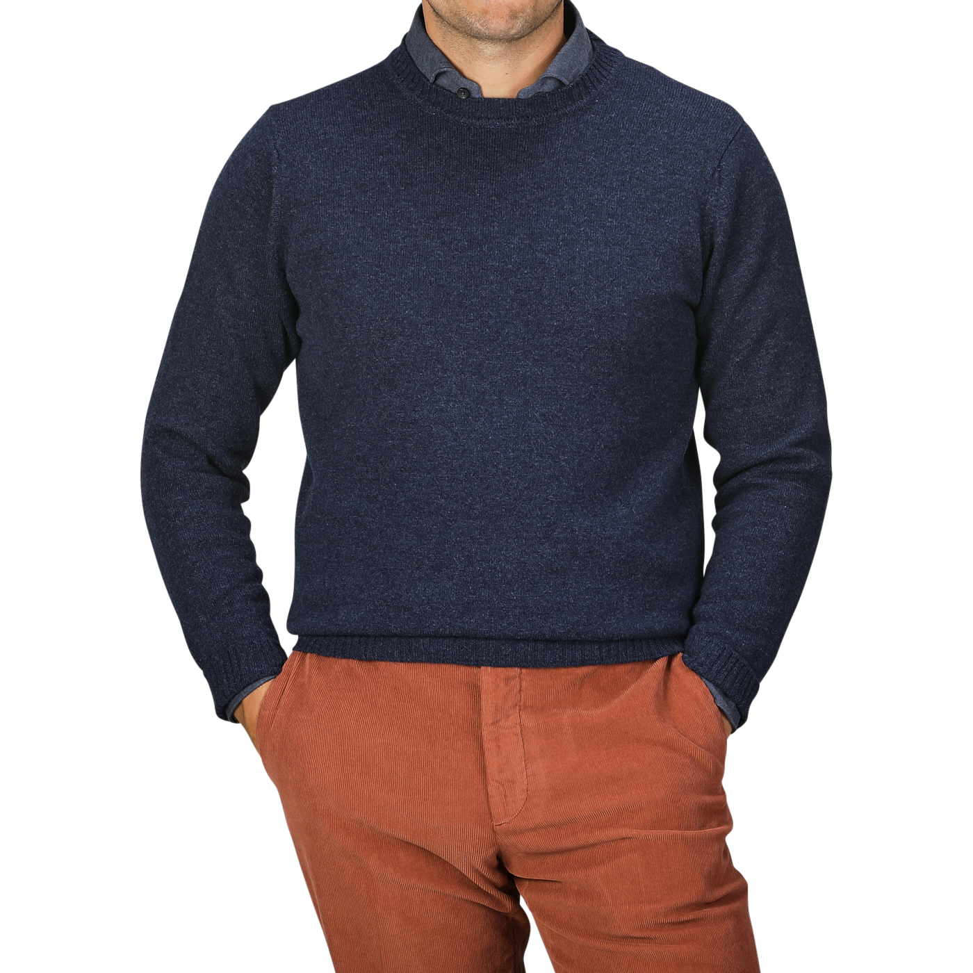 A person wearing an Astra Blue Crew Neck Lambswool Sweater by William Lockie and orange-brown pants stands with hands in pockets against a neutral background.