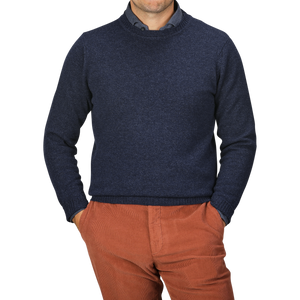 A person wearing an Astra Blue Crew Neck Lambswool Sweater by William Lockie and orange-brown pants stands with hands in pockets against a neutral background.