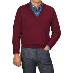 A person wearing a William Lockie Bordeaux Deep V-Neck Lambswool Sweater over a blue button-down shirt with denim jeans. The background is plain gray.