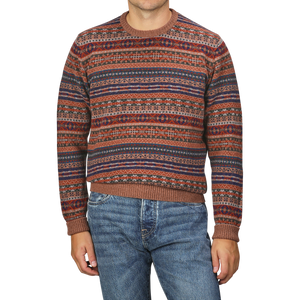 A person wearing a Brown Fair Isle Crew Neck Lambswool Sweater from William Lockie, featuring multicolored patterns, paired with blue jeans and standing against a plain background.