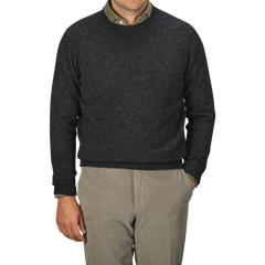 A man in a William Lockie Charcoal Grey Crew Neck Lambswool Sweater and beige pants stands against a plain background, with his left hand partially visible in his pocket. Only his torso is shown.