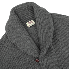 The Cliff Grey Lambswool Shawl Collar Cardigan from William Lockie is showcased. Made from soft Scottish lambswool, this gray, ribbed-knit cardigan features a single maroon button near the bottom of the collar.