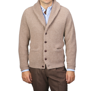 A person is dressed in a Colt Beige Lambswool Shawl Collar Cardigan by William Lockie over a light blue shirt and brown pants. The cardigan, adorned with leather buttons and two front pockets, adds a touch of timeless elegance.