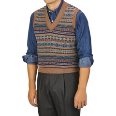 A person wearing a William Lockie Driftwood Brown Fair Isle V-Neck Lambswool Slipover over a denim shirt with rolled-up sleeves and dark trousers.