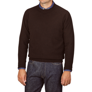 A person wearing a William Lockie Ebony Brown Crew-Neck Lambswool Sweater over a blue shirt with jeans, standing against a plain background.