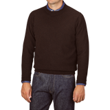 A person wearing a William Lockie Ebony Brown Crew-Neck Lambswool Sweater over a blue shirt with jeans, standing against a plain background.