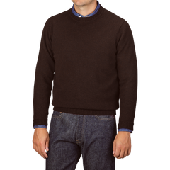 A person wearing a William Lockie Ebony Brown Crew-Neck Lambswool Sweater over a blue shirt with jeans, standing against a plain background.